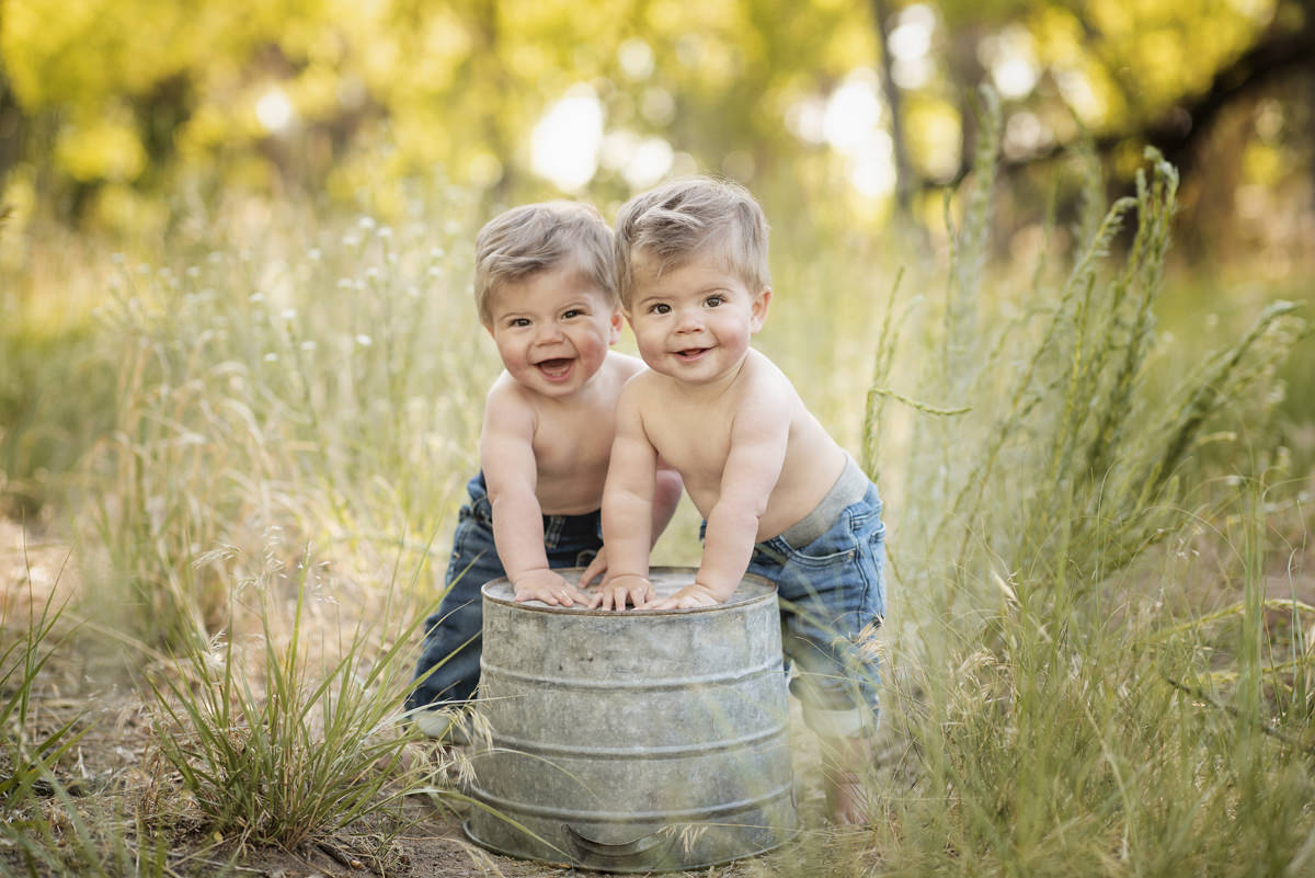 One year old boy twins in jeans with their shirt off supporting themselves on a bucket before they can walk
