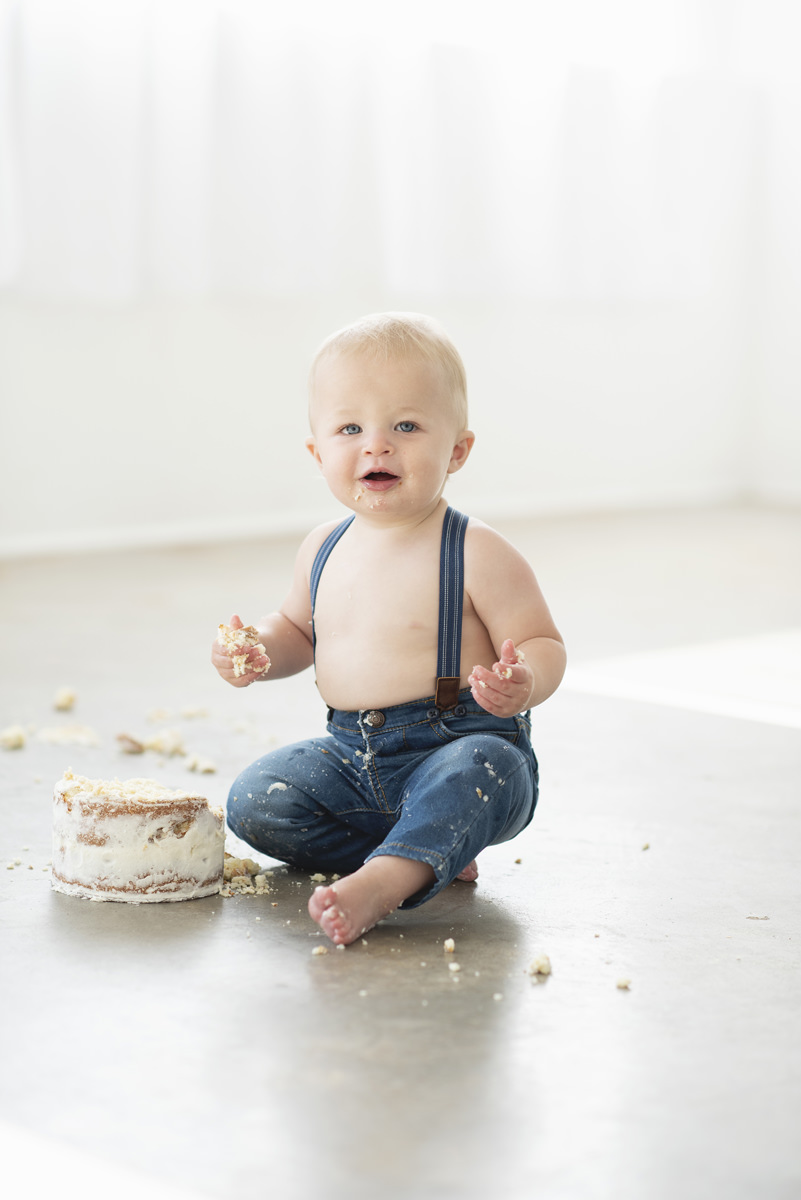 Baby sitting on a cement floor in jeans and suspenders with his shirt off eating a smash cake to celebrate his first birthday