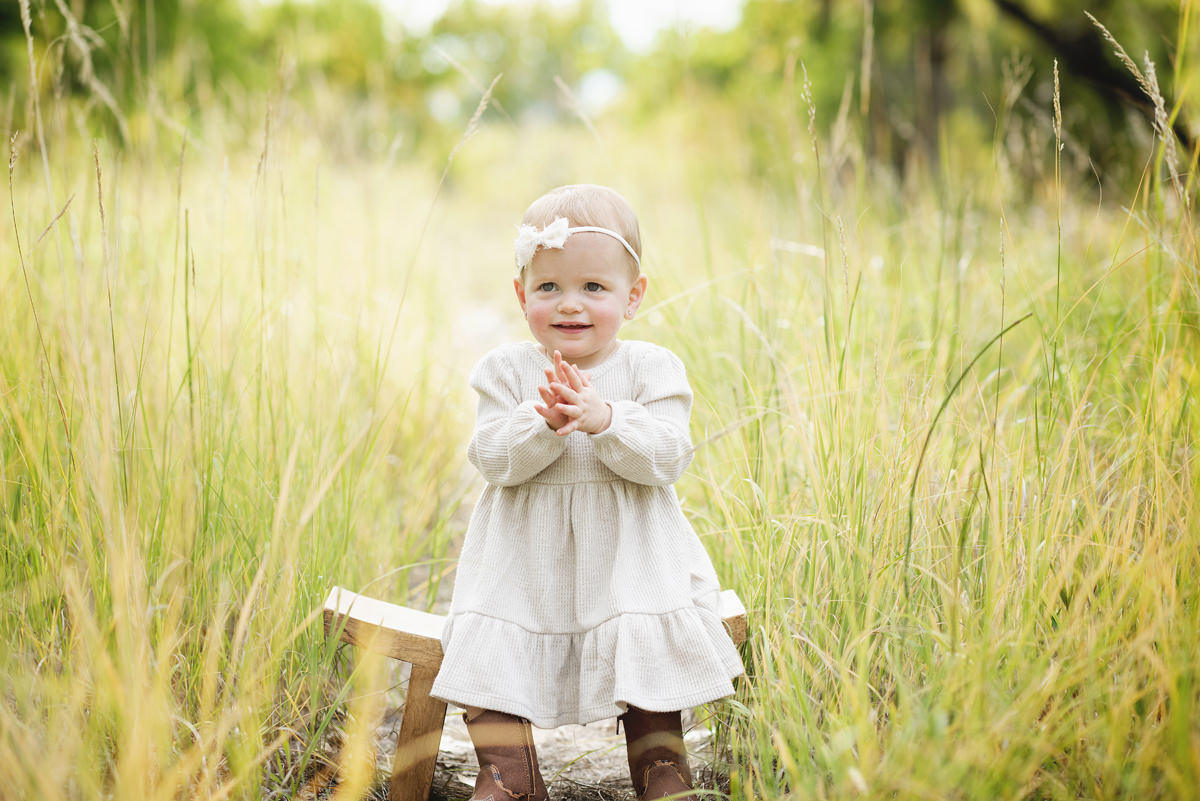 18 month old toddler sitting on a small wooden bench in long grass with her hands clapping and wearing a neutral colored dress