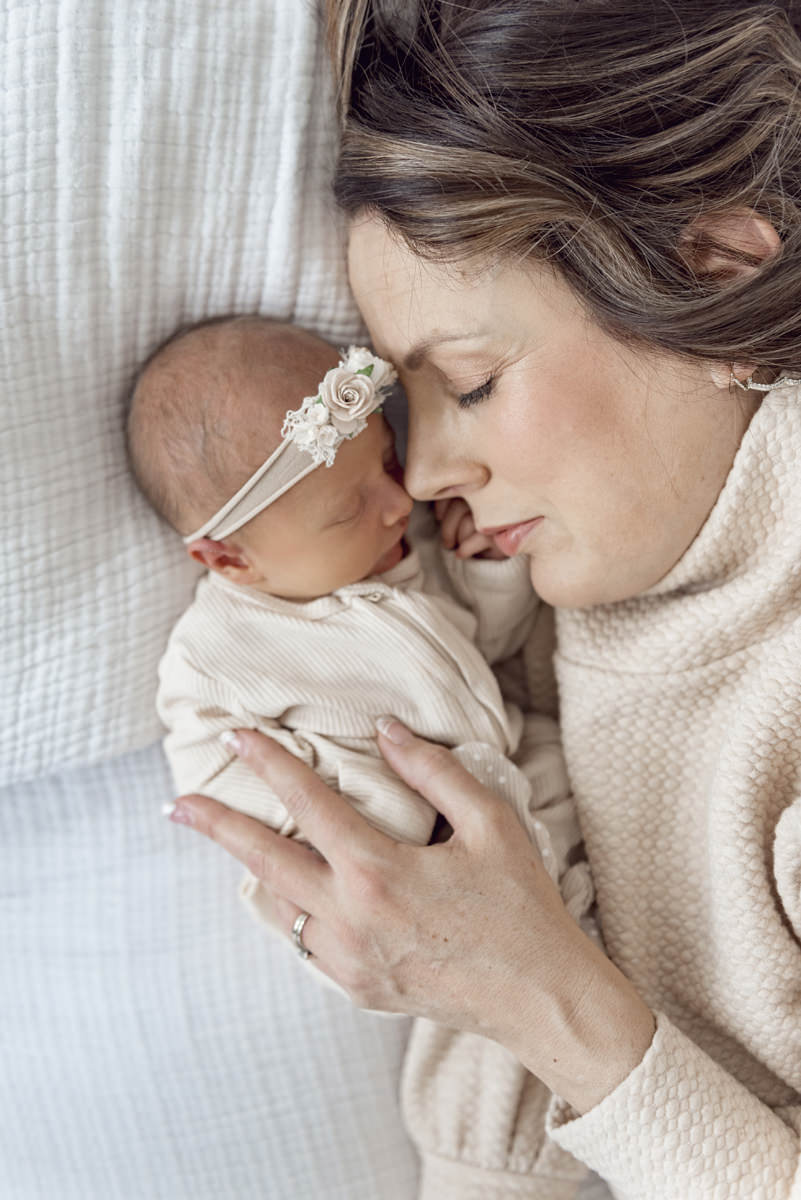 Newborn baby and mom snuggling on a bed with a white comforter, with baby and mom dressed in a peach color