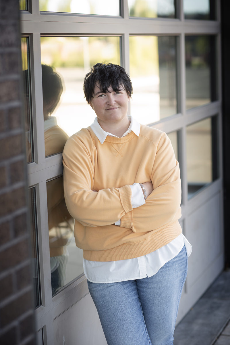 High school senior girl with short hair standing in front of a garage door with windows wearing a canary yellow sweater with a white collared shirt underneath in 2023