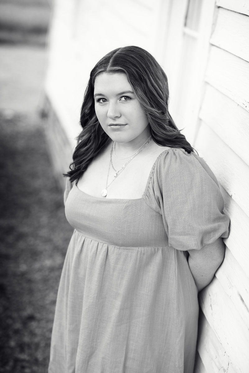 black and white high school senior girl in a dress leaning against the side of a white farmhouse in Parker, Colorado