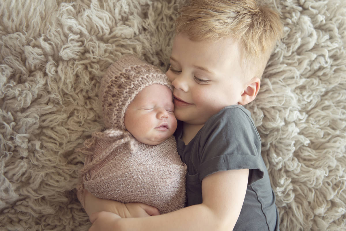 Sibling newborn photography, brother hugging baby sister who is wearing a pink bonnet and matching wrap