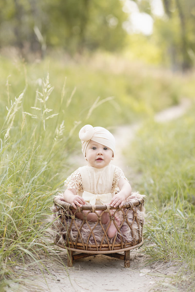 7 month old baby sitting in a boho basket in outdoors in long grass along a dirt path wearing a knot head turbine dressed in lacy off white in the summer