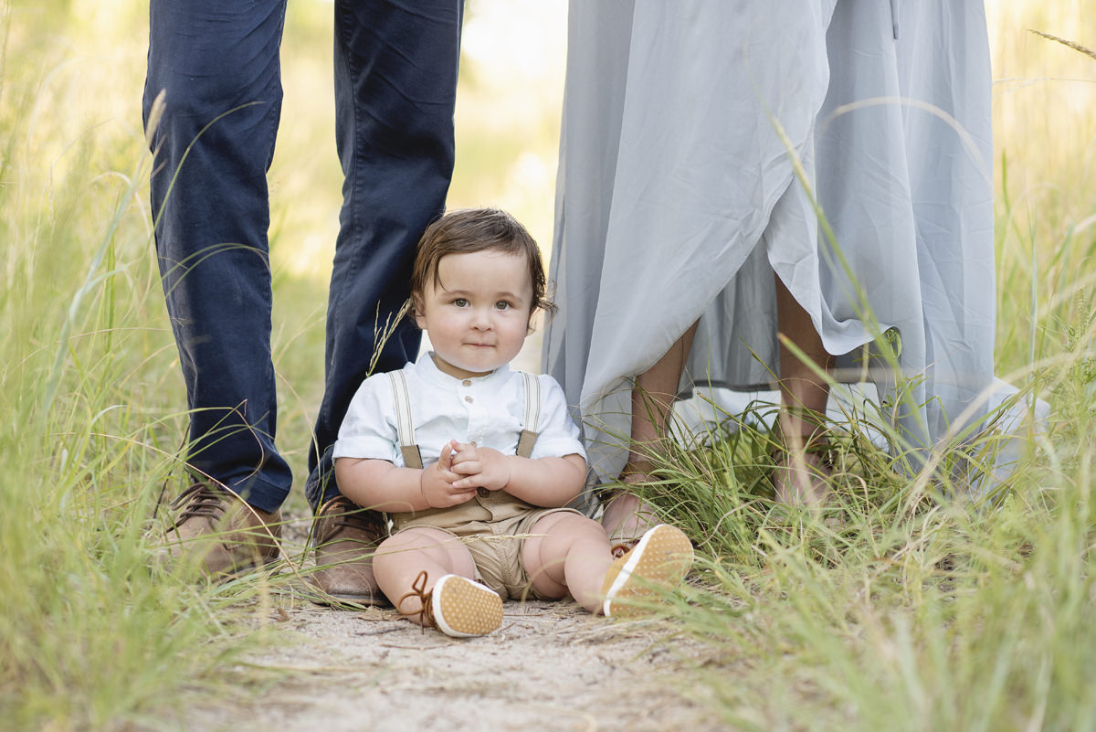 one year old baby boy sitting in front of his parents feet outdoors in the long wild grass