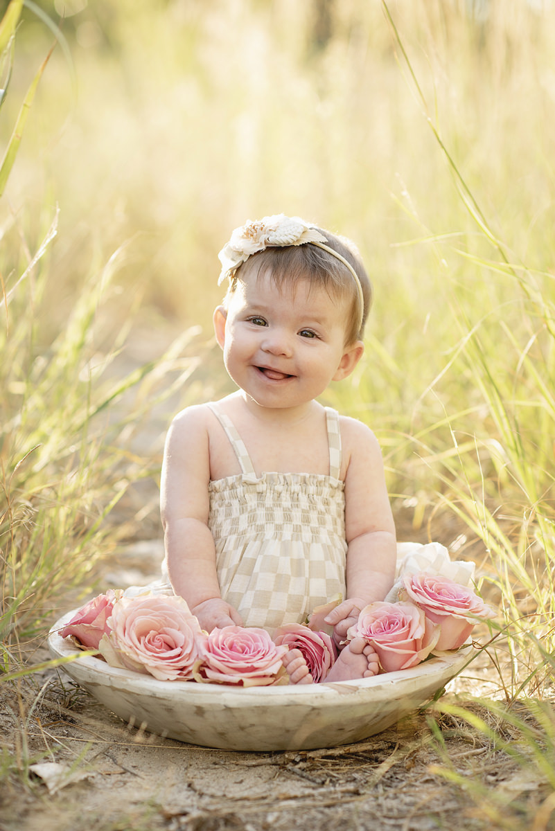 8 month old baby girl sitting in a bowl outdoors in the golden sunlight with tall grass and pink roses in the bowl with a headband and tongue sticking out