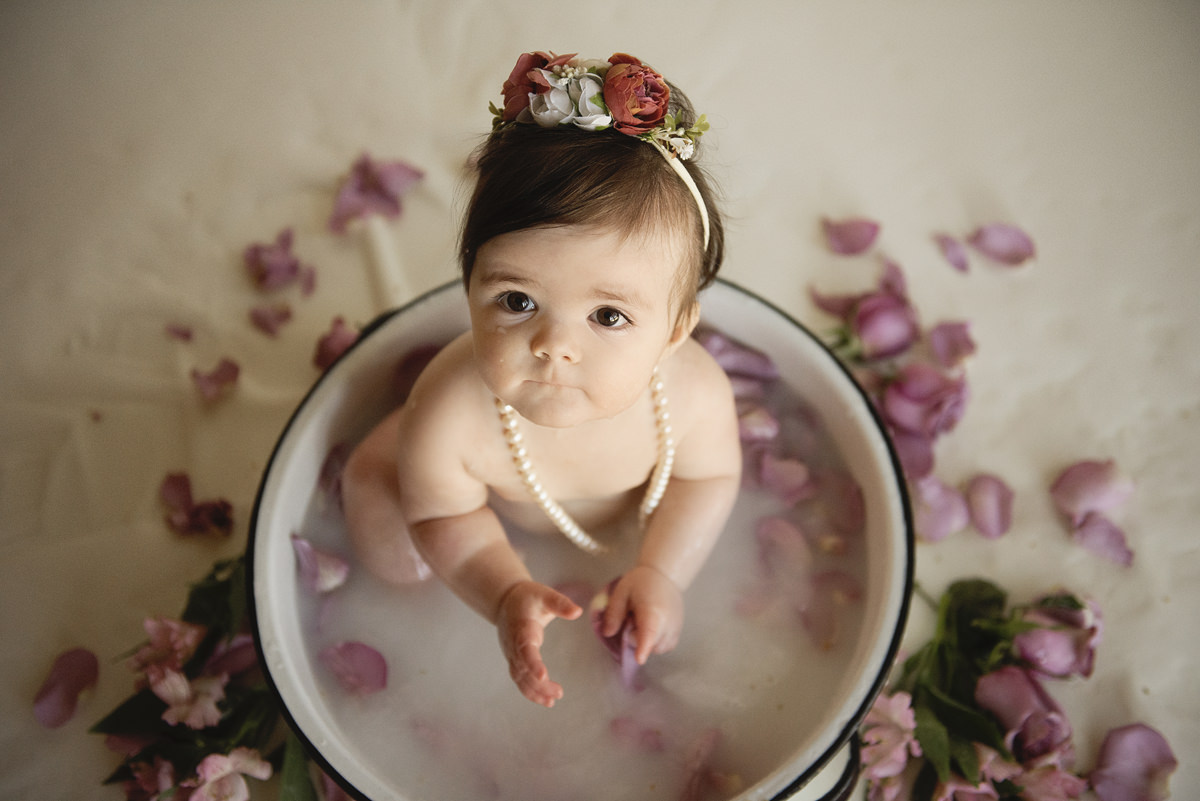 IN-Home photography session against a white backdrop of a one year old baby in a white metal bucket milk bath with whole roses, rose pedals, and other flowers