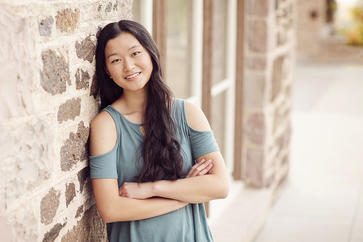 Senior girl leaning against a stone wall wearing a teal blue shirt; Parker senior pictures review