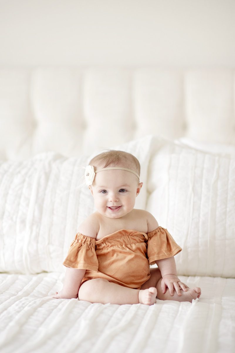 Gorgeous professional photography of a six month old baby girl sitting on a white bed wearing a rust colored off the shoulder one piece taken by Parker Milestones Photographer Jamie Herrera