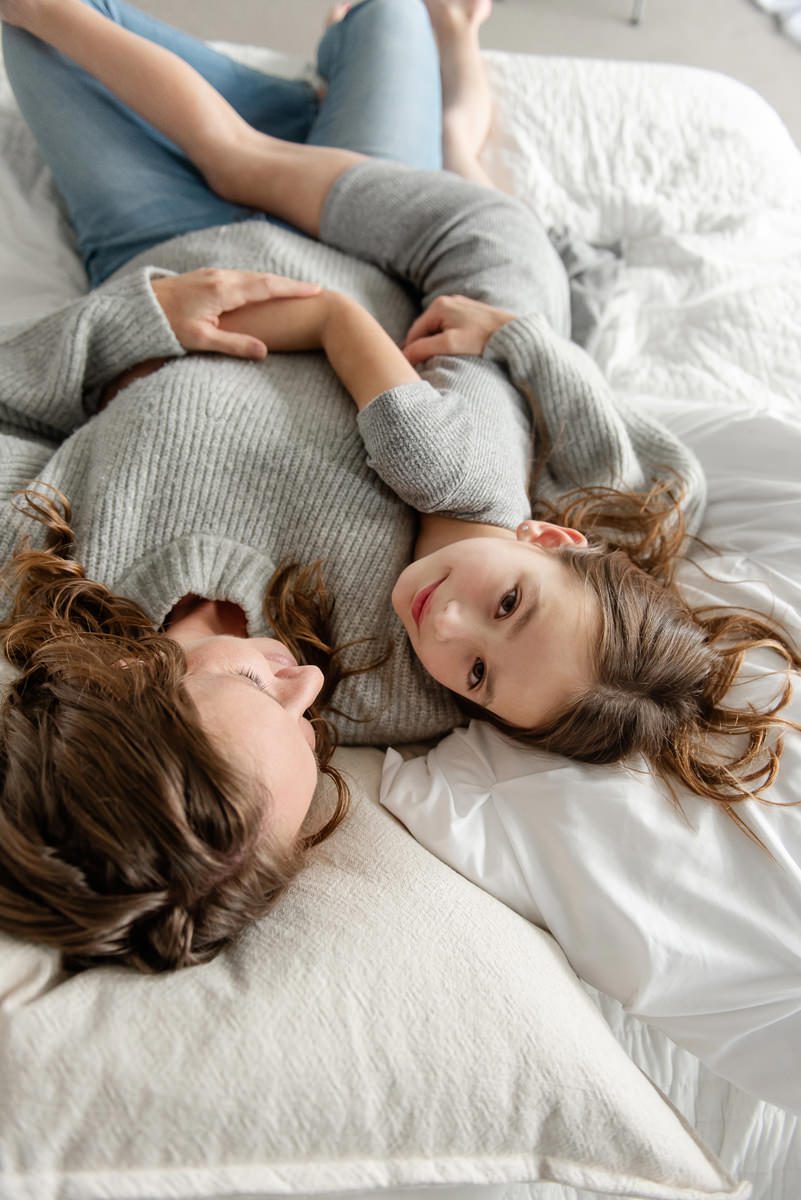 picture of a girl looking at camera laying in her mom's arms on a bed taken by Parker Family Photographer, Jamie Herrera