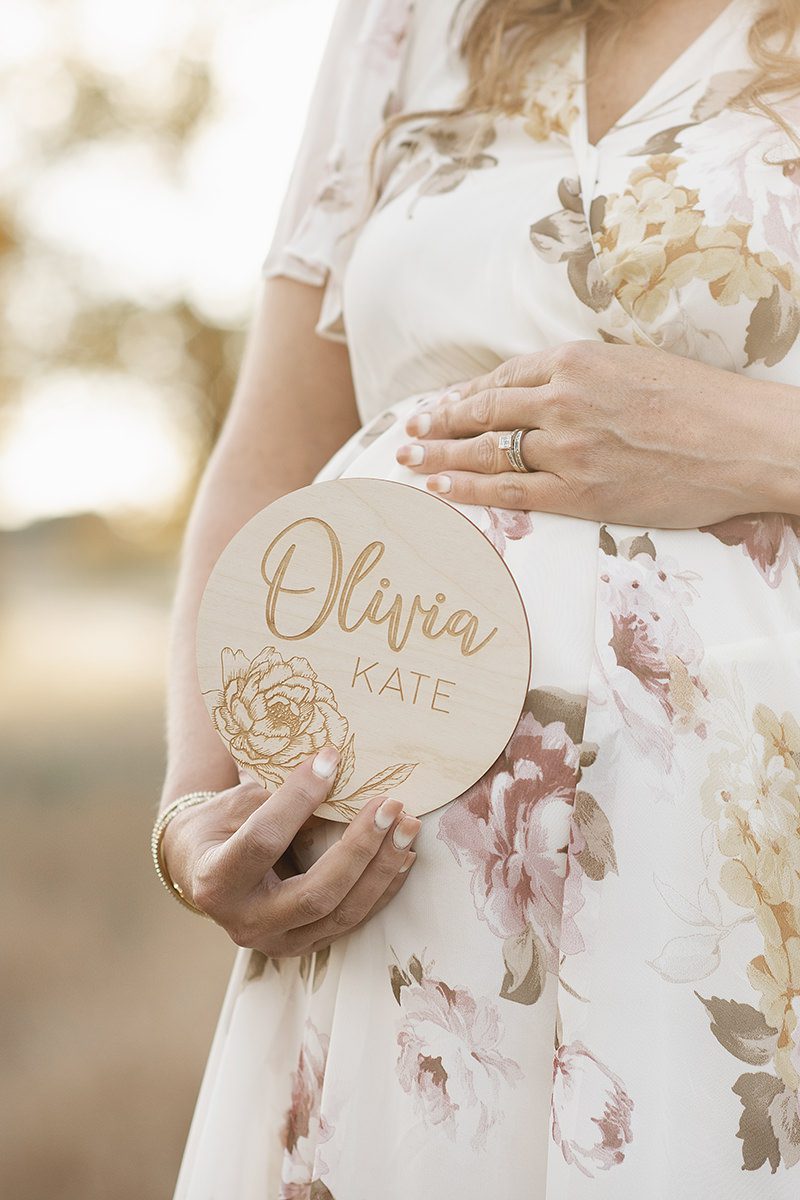 pregnant mom, close up shot of her belly holding. a round wood sign with the baby's name on it