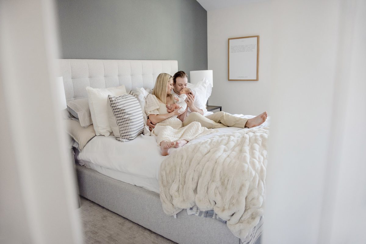 aurora newborn photographer capturing parents sitting on a bed holding their baby in a bright, cozy in-home lifestyle setting