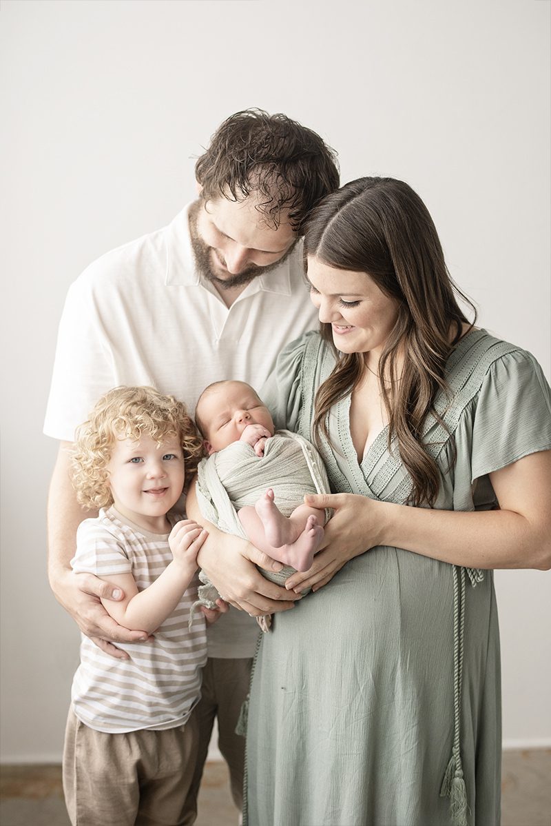 aurora newborn photography session with parents holding their swaddled baby while a curly-haired toddler stands close, soft neutral studio setting