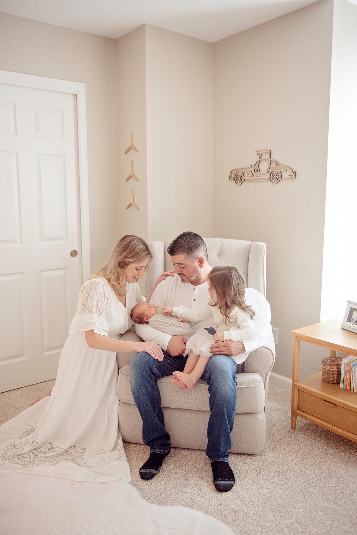 family gathered around their newborn in a cozy nursery, parents seated with baby while sibling leans in close, soft natural light and neutral tones