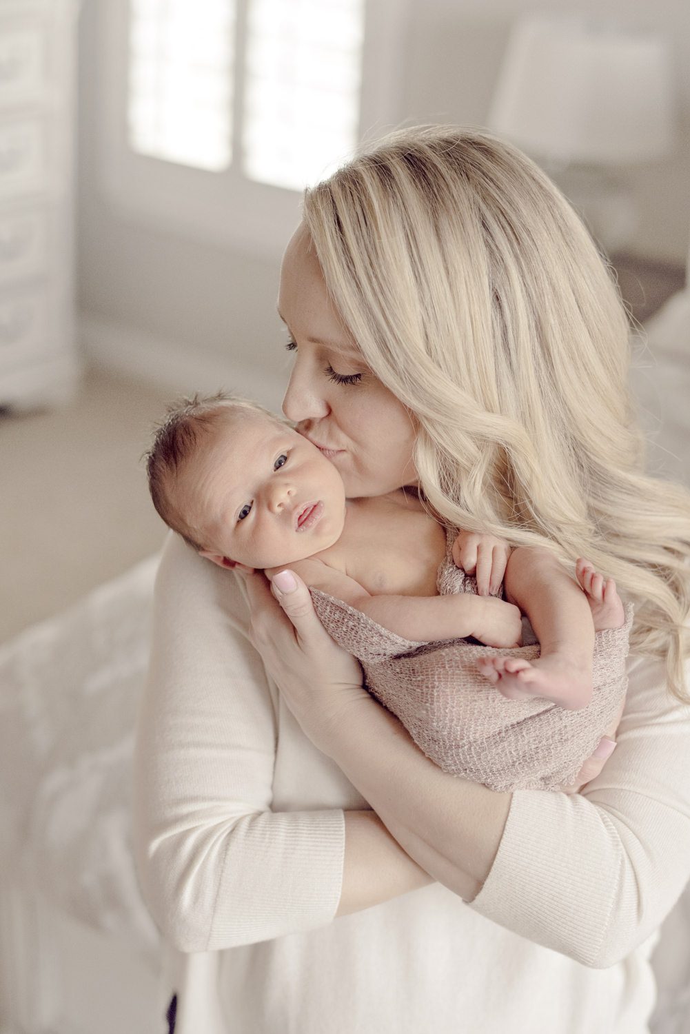 mother holding her newborn close and kissing the baby’s cheek, soft window light and calm neutral tones